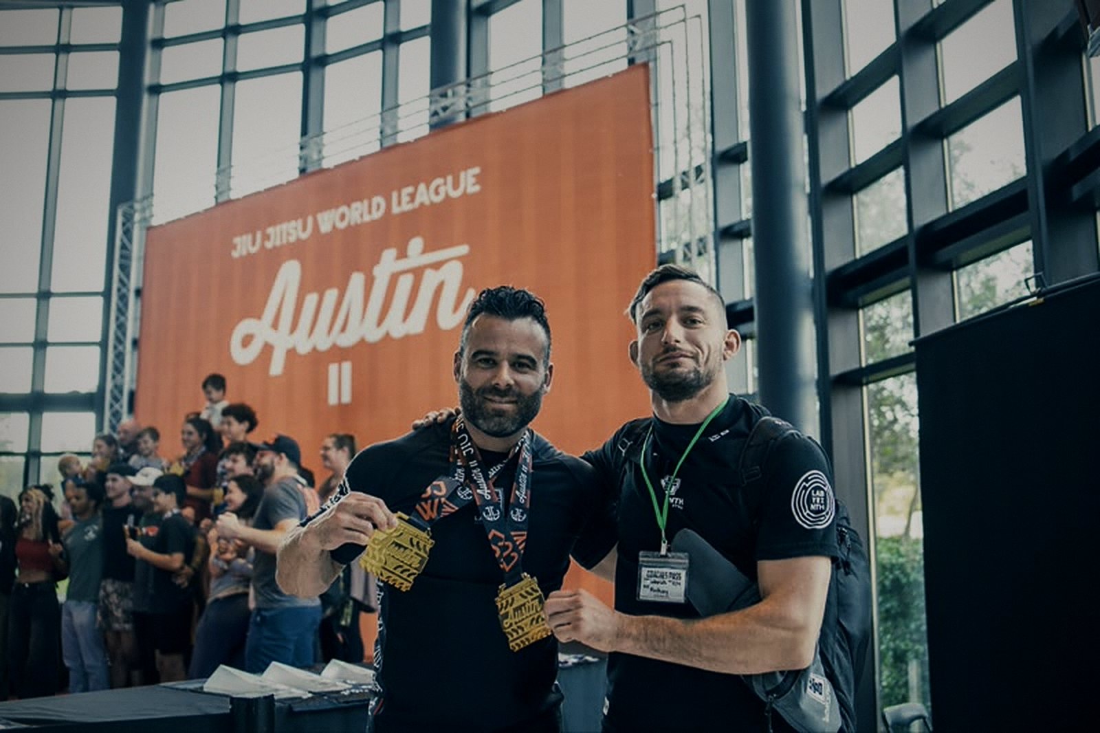 Two Labyrinth BJJ team members hold gold medals in front of the Jiu Jitsu World League Austin banner