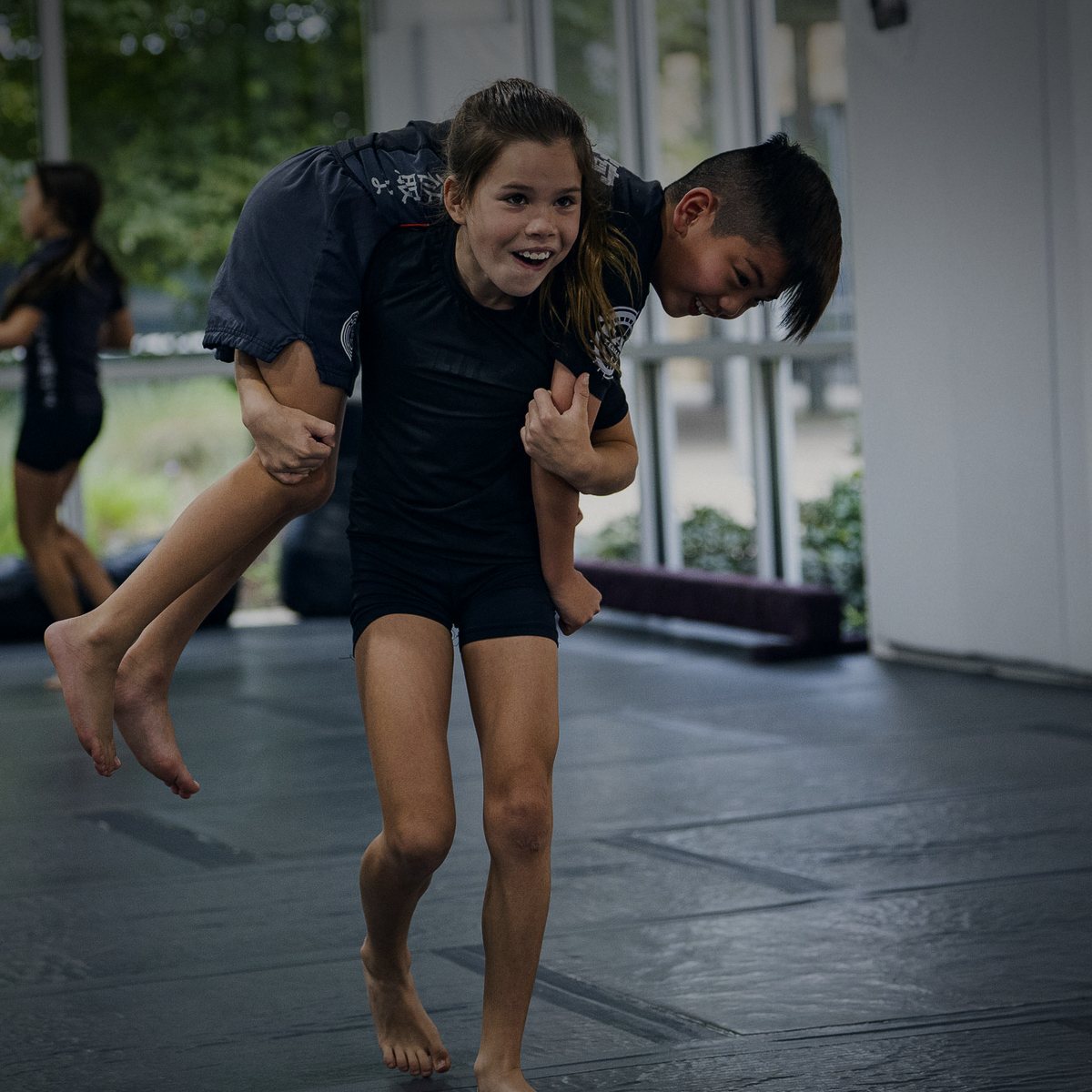 A smiling kid carries another kid across the shoulders in a kids class at Labyrinth BJJ in Fulshear, TX